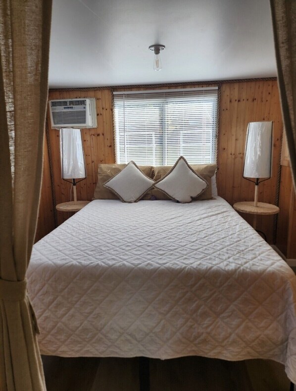 A bedroom with a bed, two bedside tables, a window with blinds, and an air conditioning unit.