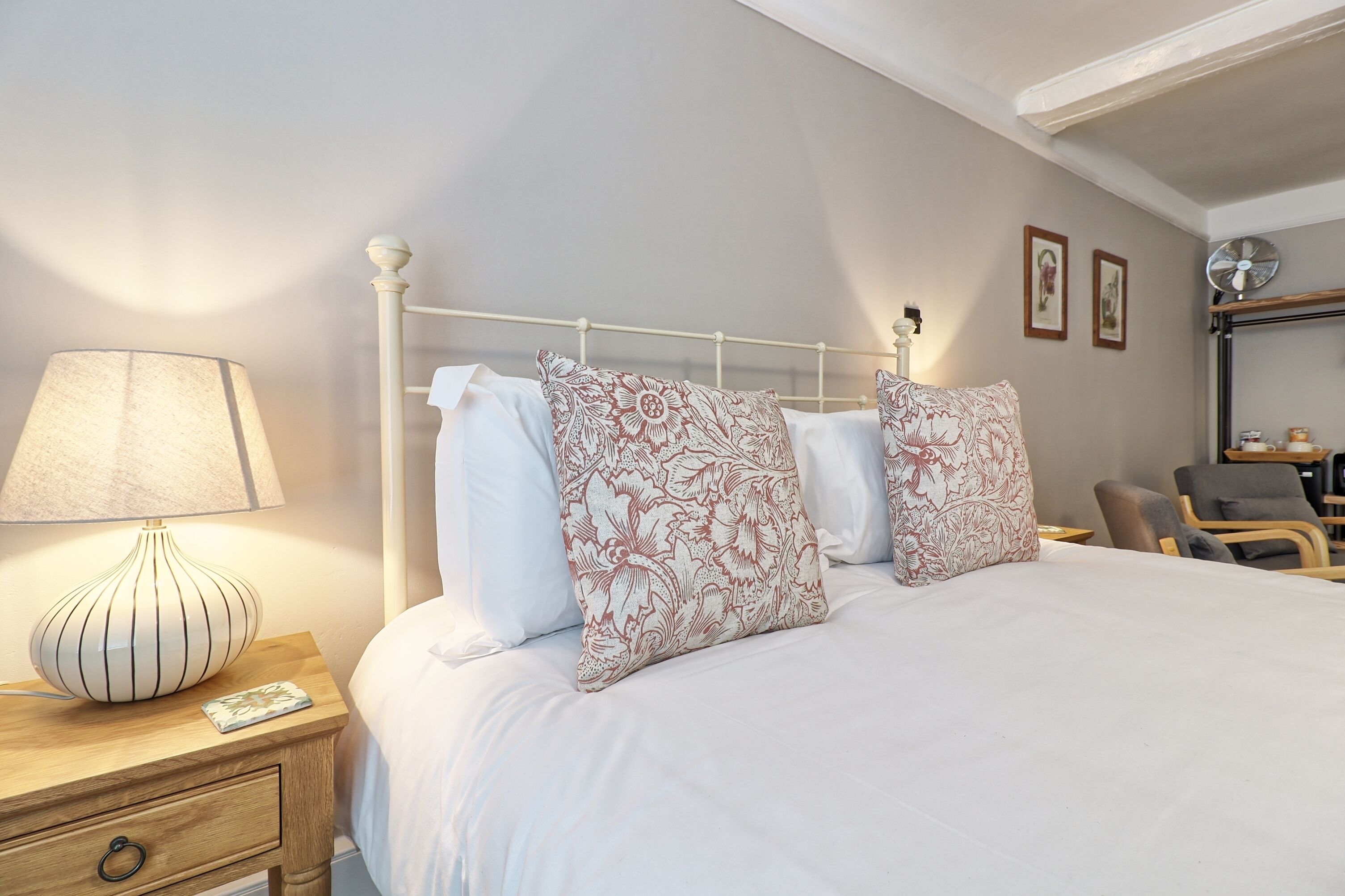A neatly made bed with floral-patterned pillows, a bedside lamp, and a chair in a well-lit room with a simple, clean design.
