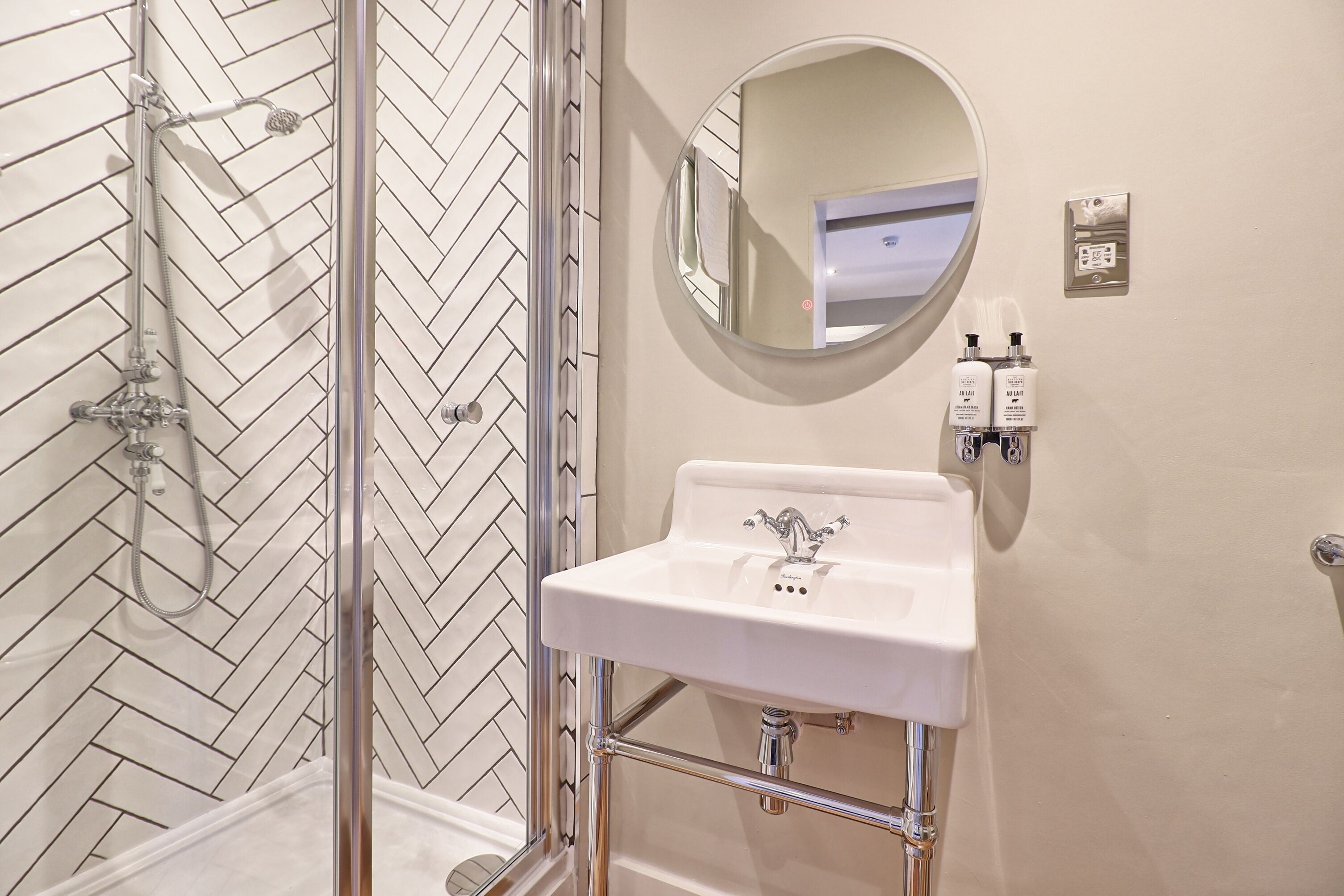 Une salle de bain avec un mur recouvert de carreaux disposés en motif «chevrons», une cabine de douche en verre, un lavabo blanc avec des robinets chromés, un miroir rond et deux flacons de produits de toilette fixés au mur.