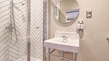 Een badkamer met een backsplash van mozaïektegels in een herringbone patroon, een glazen douchecabine, een wit wastafelmeubel met chroombeslag, een ronde spiegel en twee toiletartikelen aan de muur.