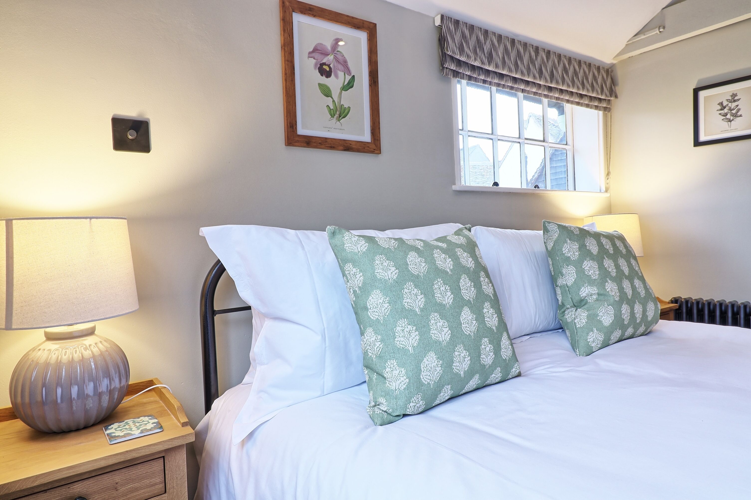 A neatly made bed with white and green patterned pillows, a bedside lamp, a framed botanical print on the wall, and a window with a valance.