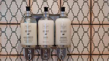 Trois flacons de produits de bain de la marque « Au Lait » (shampoing, après-shampoing et gel douche) sont fixés sur un mur orné d’un motif géométrique en carreaux.