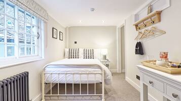 A bedroom with a bed, a bedside table, a radiator, a window with curtains, a wall-mounted shelf with hangers, and a tray with items.