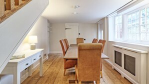 A dining area with a wooden table and chairs, a white console table with a lamp, a staircase, and a window with a view. - Noble Goose Rooms (Colchester)