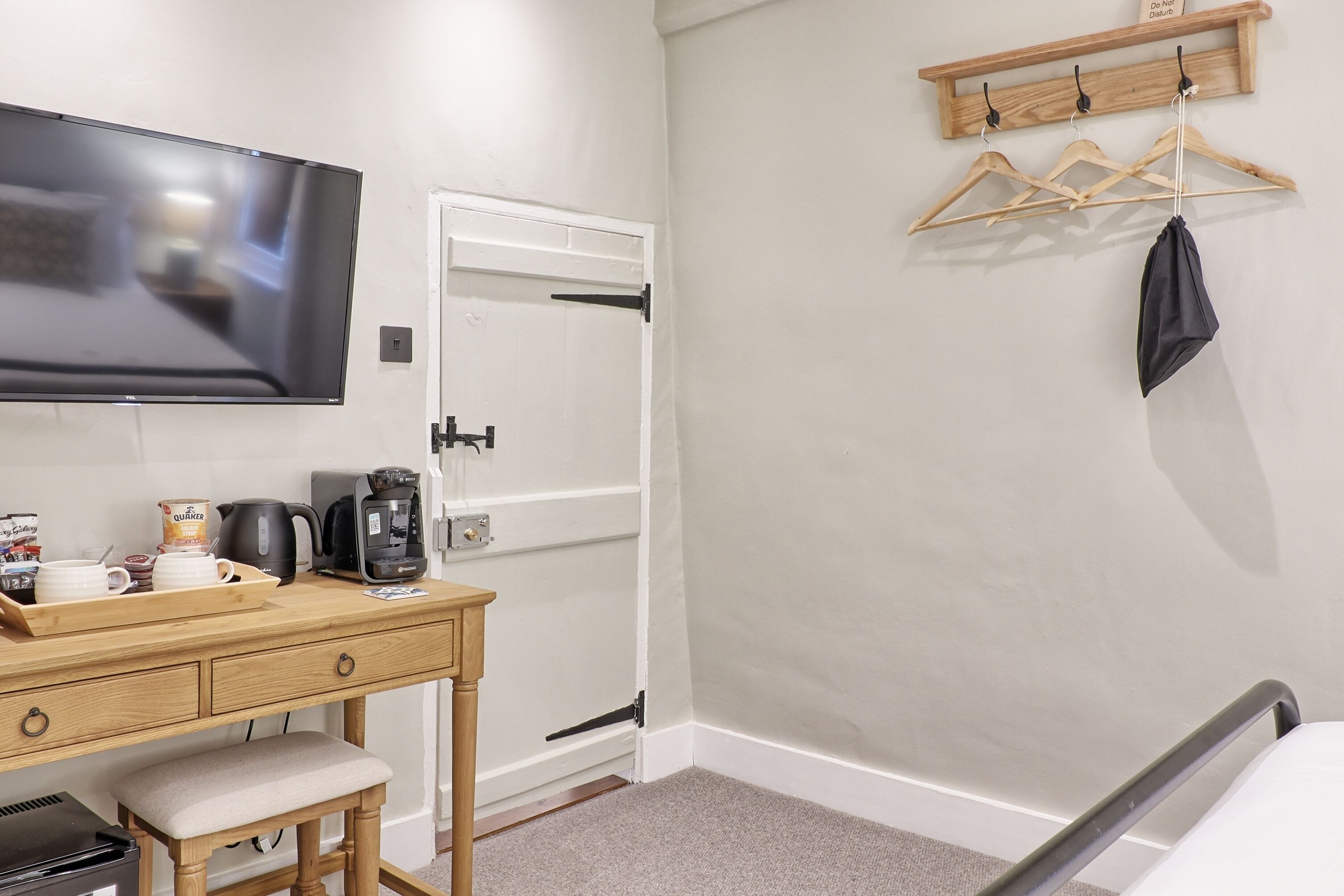 Habitación de hotel con televisor de pantalla plana, un escritorio de madera con cafetera, hervidor y una bandeja con productos para el desayuno. Se observa una cama con cabecera y un perchero de madera con perchas.