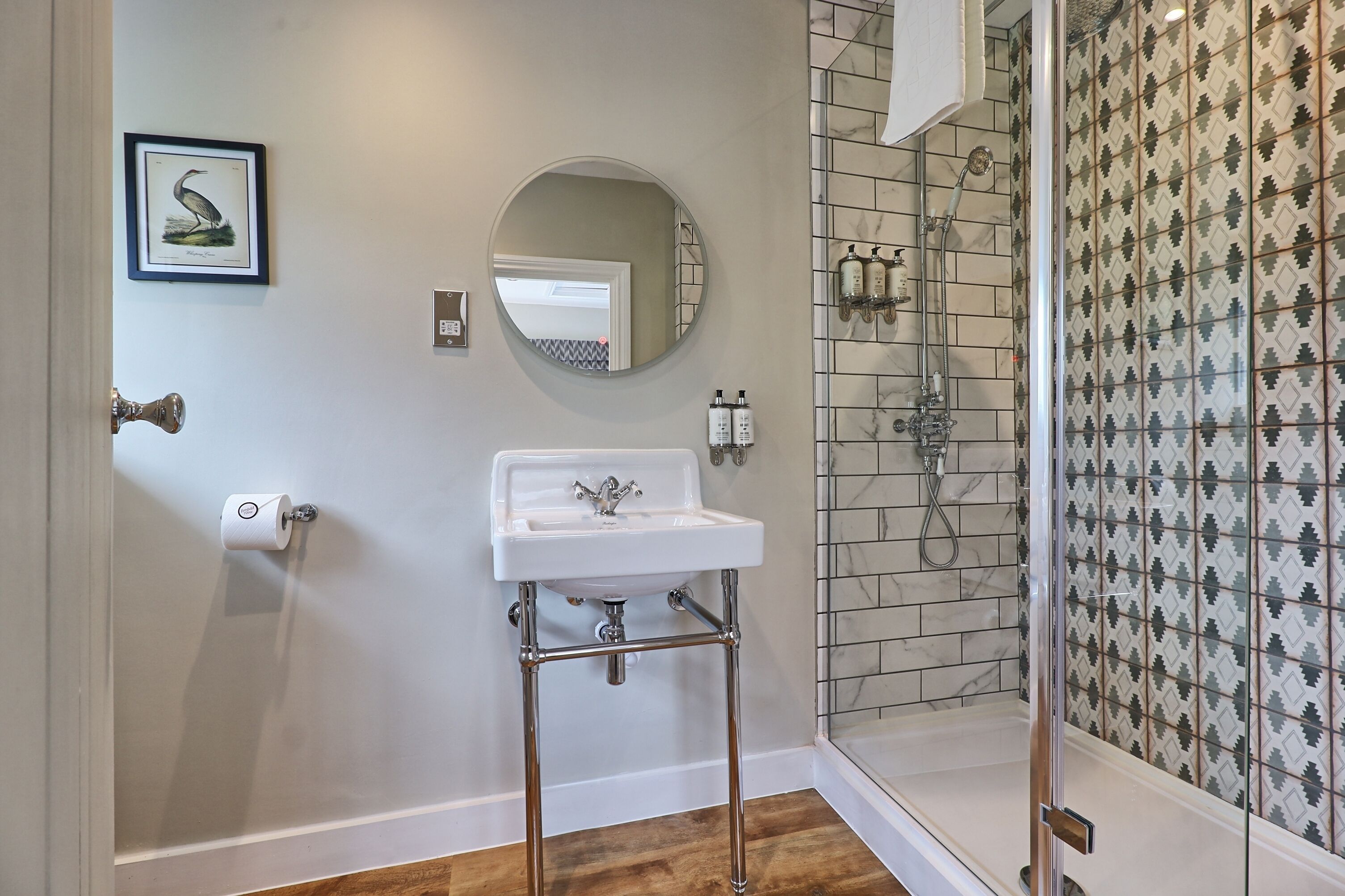 A bathroom with a white sink, a round mirror, a shower with a glass door, and patterned tiles on the walls.