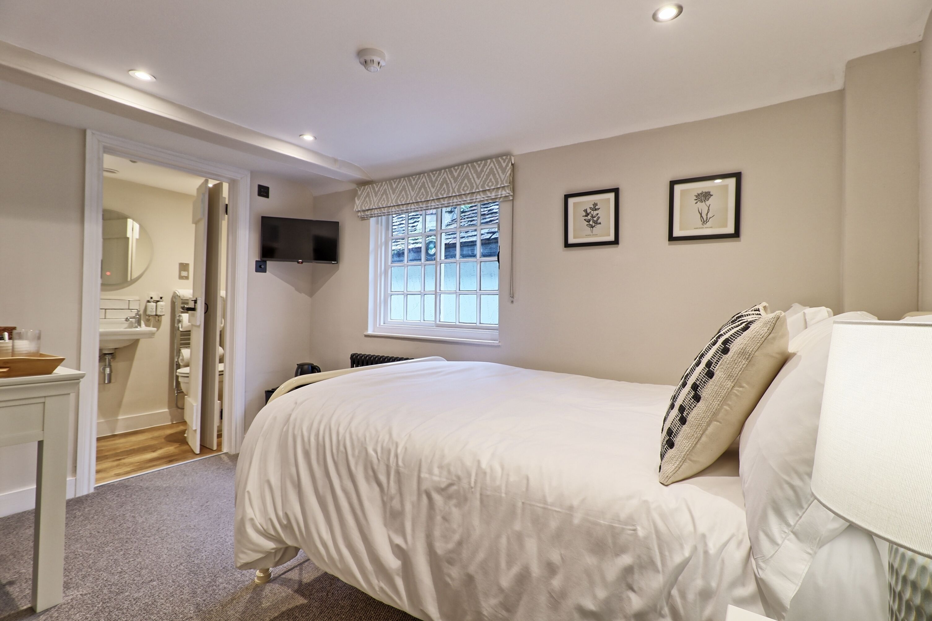 A bedroom with a bed, a window with curtains, a TV mounted on the wall, and a bathroom visible through an open door.