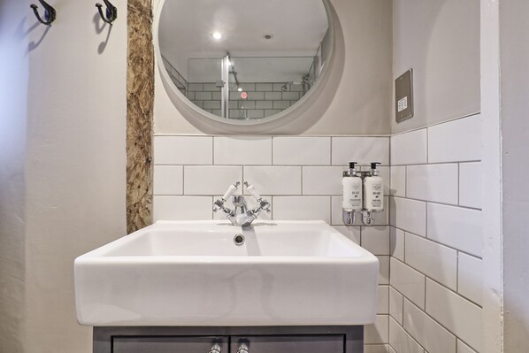 A bathroom with a white sink, a round mirror, and a shower visible in the reflection. - Noble Goose Rooms (Colchester)