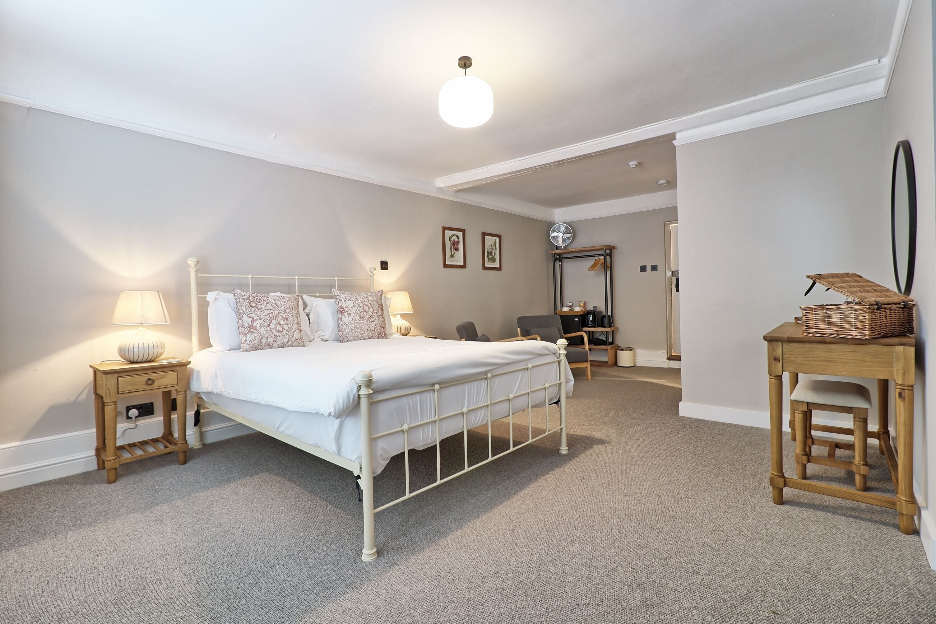 Un dormitorio con una cama de metal blanco, dos mesitas de noche con lámparas, una silla de madera, una pequeña mesa con una cesta y un espejo en la pared.