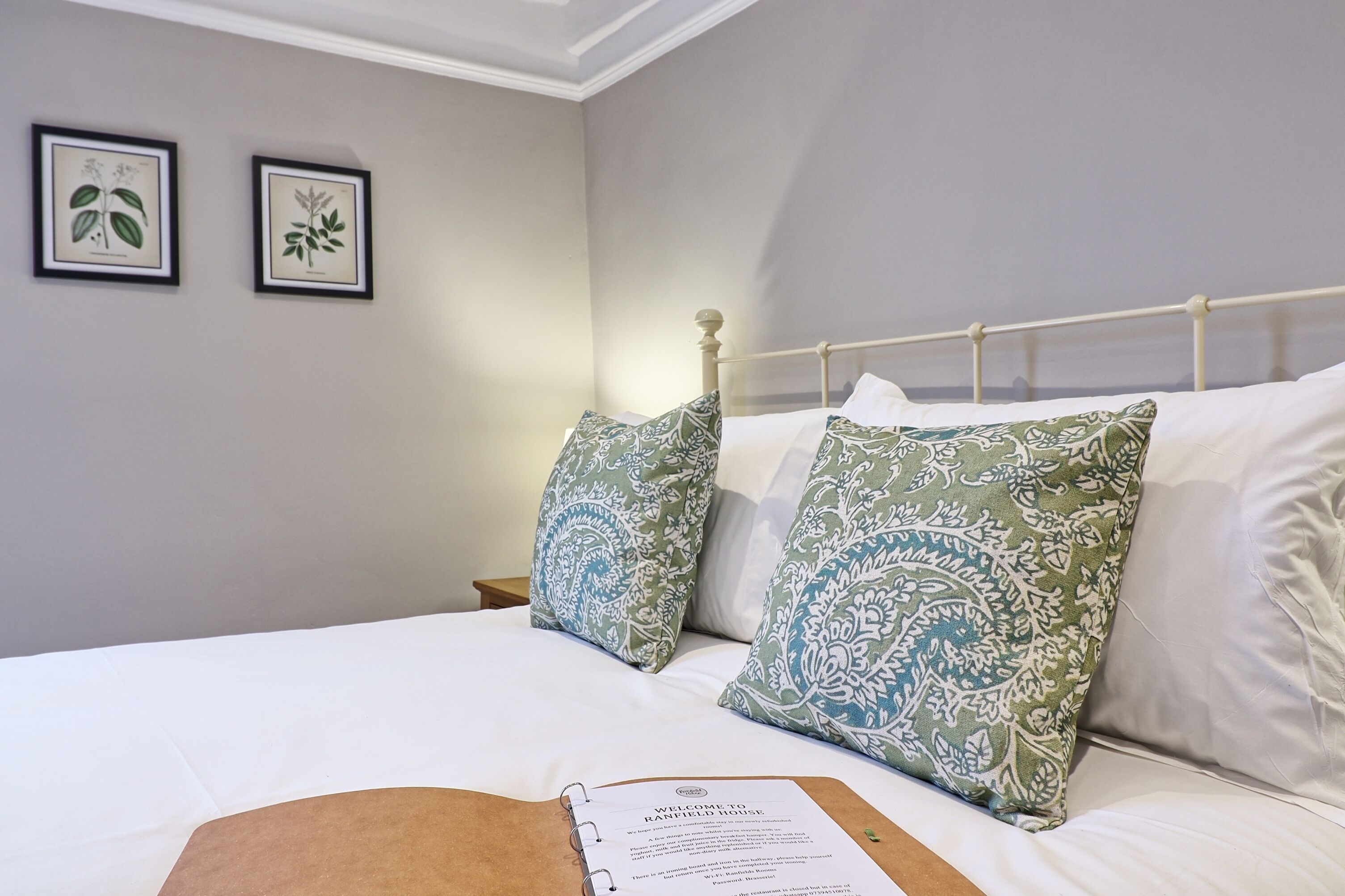 A neatly made bed with patterned pillows, a welcome booklet, and botanical wall art.