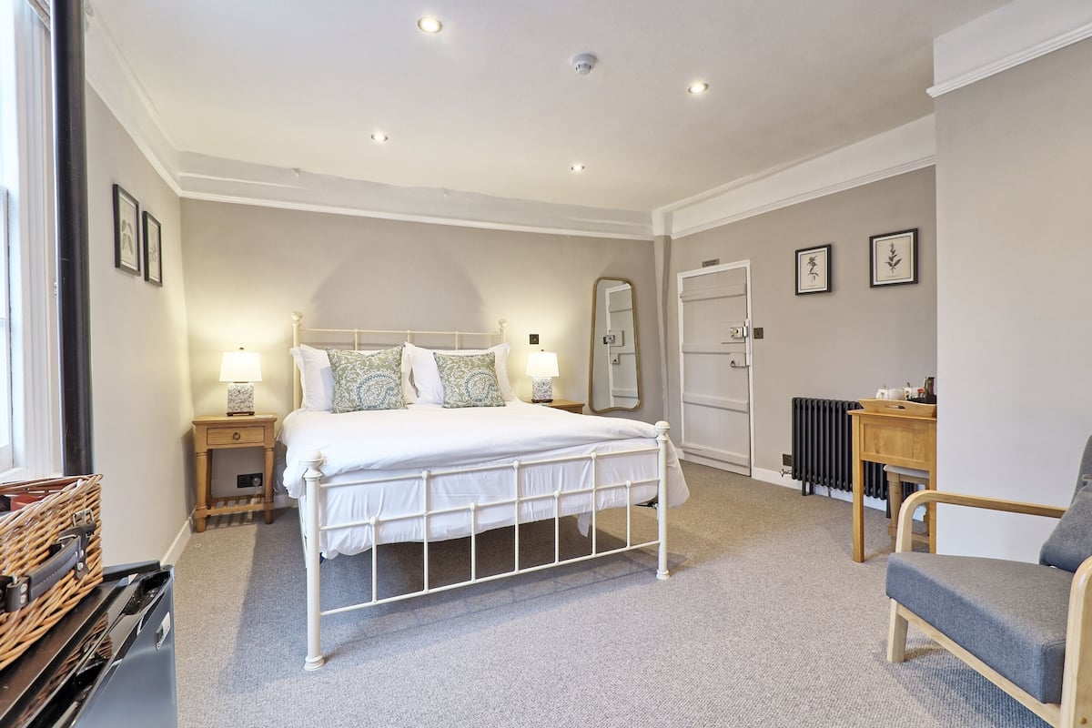 A bedroom with a white metal bed, two bedside tables with lamps, a wooden chair, a radiator, and framed pictures on the walls.