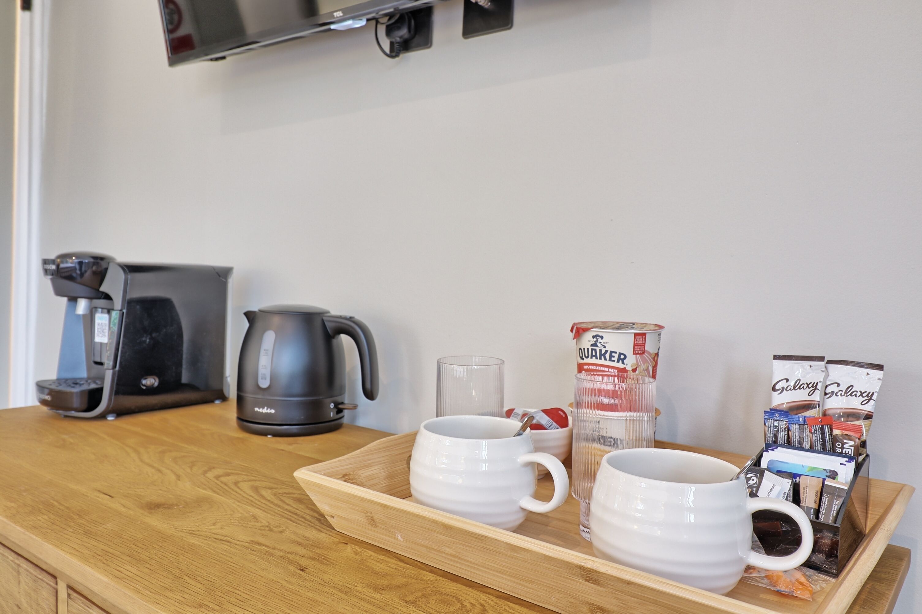 A wooden tray with two white mugs, a glass, a coffee maker, a kettle, a canister labeled 'Quaker,' and a coffee pod holder.