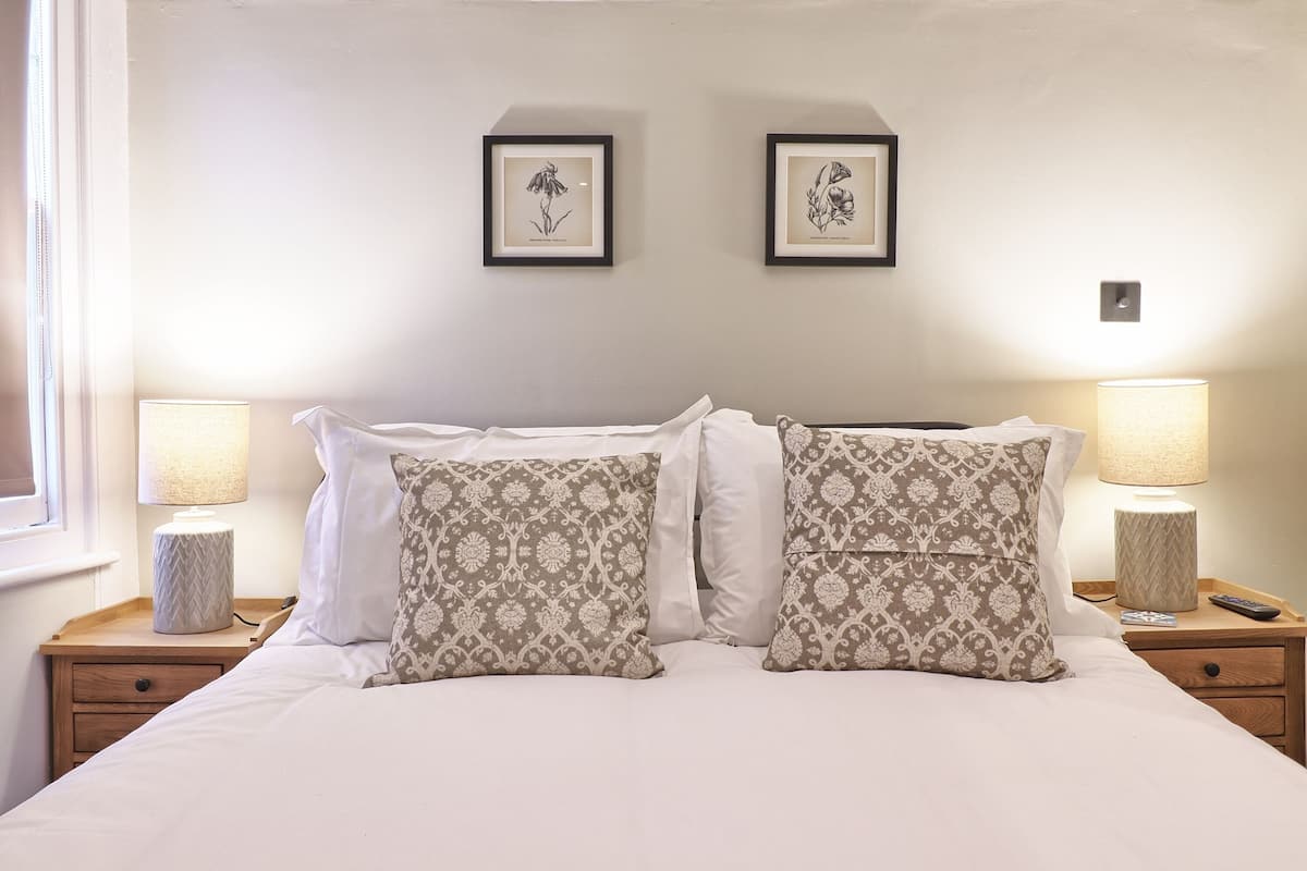 A neatly made bed with patterned pillows, two framed botanical prints on the wall, and bedside lamps on wooden nightstands.