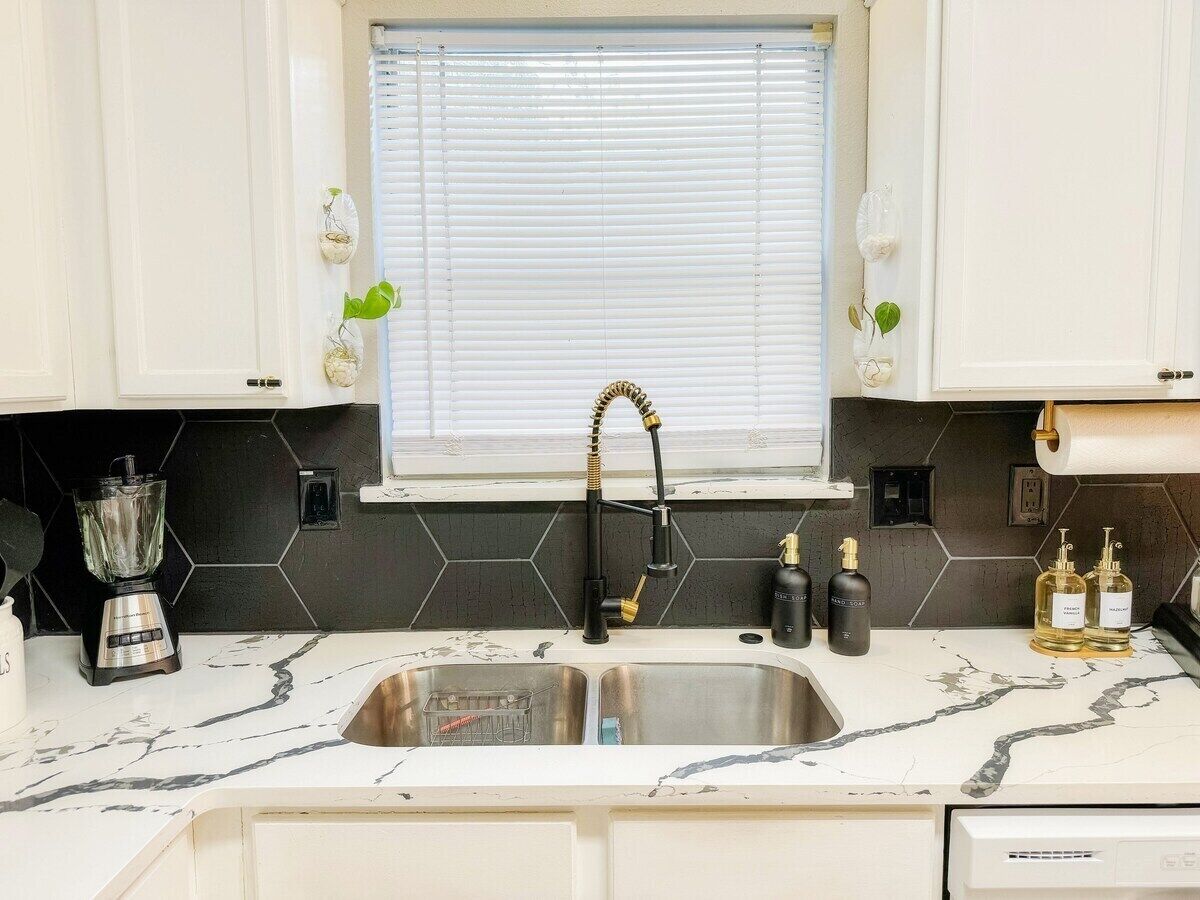 Una cocina con un lavamanos doble, grifo negro y una ventana con persianas. Hay dos botellas en la encimera y una licuadora.