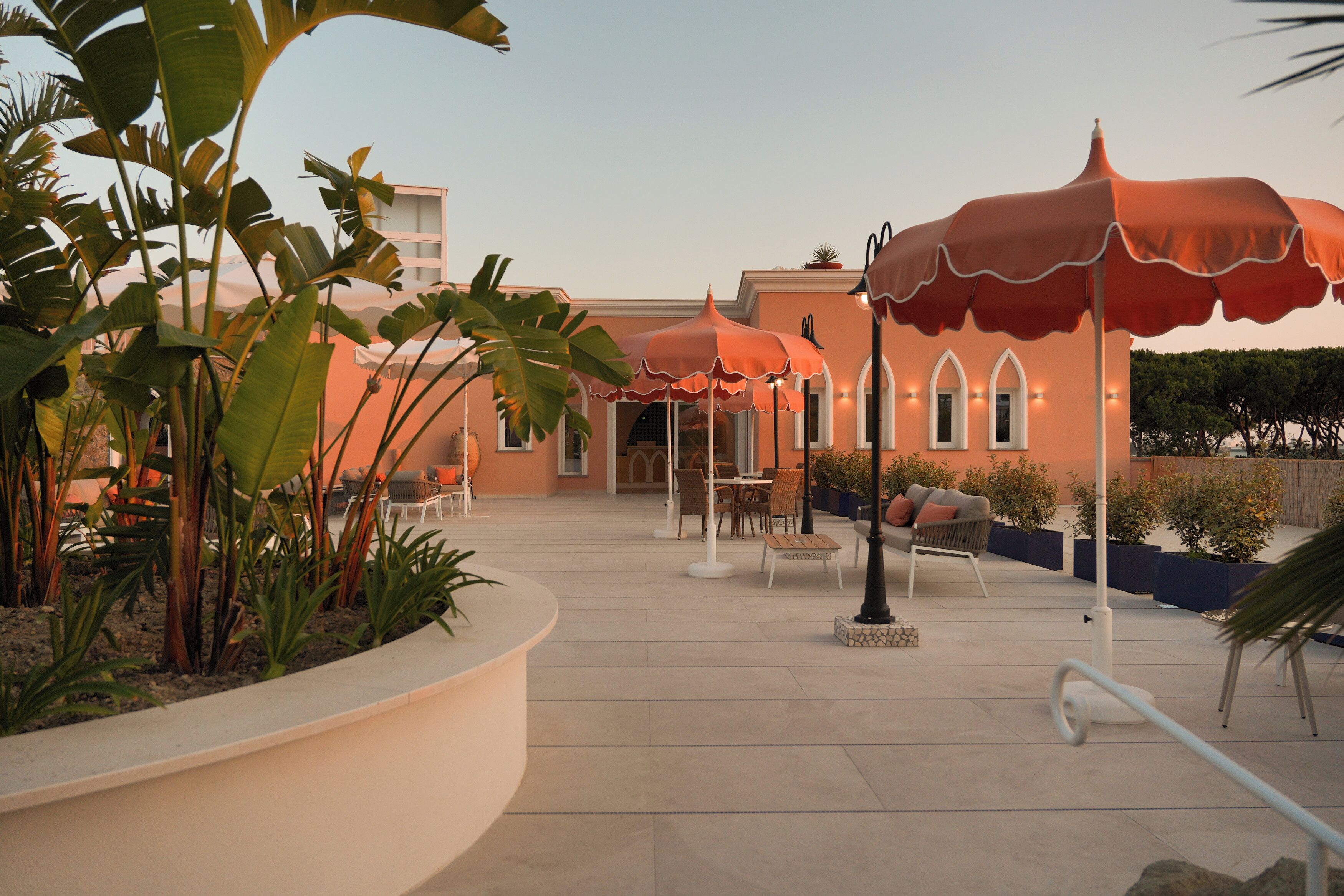 An outdoor patio area with seating and umbrellas, surrounded by plants and a building with arched windows.