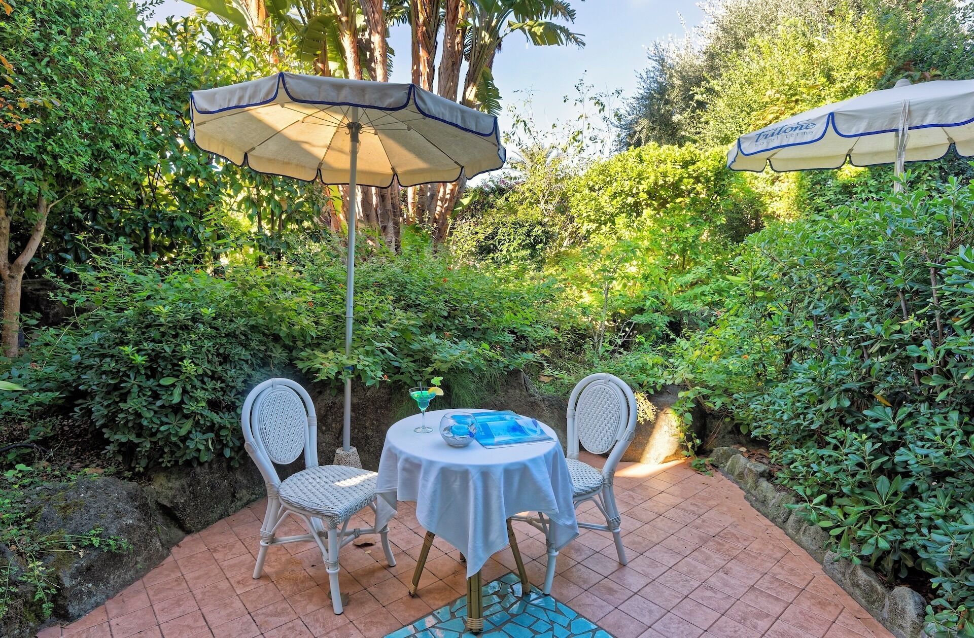 Een terras met twee witte rieten stoelen en een tafel, overdekt door een beige parasol.