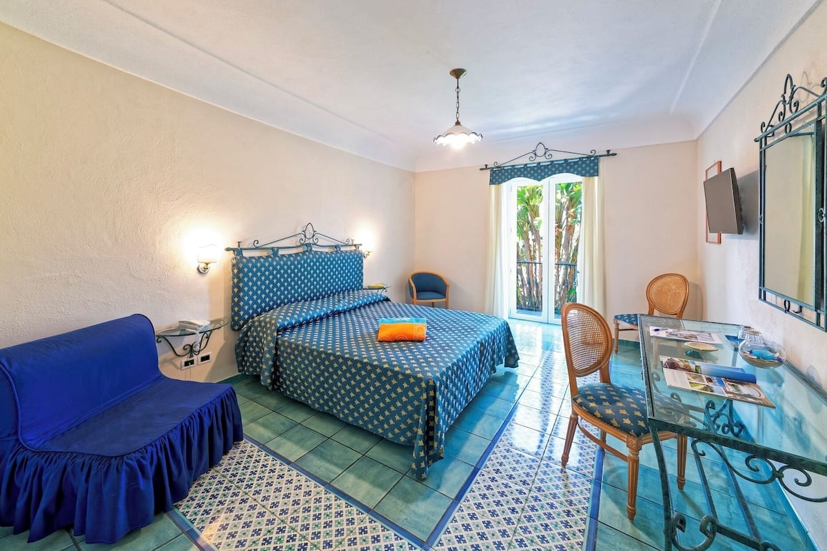 Un dormitorio con una cama, un sofá azul, una mesa de vidrio con sillas y vistas a zonas verdes.