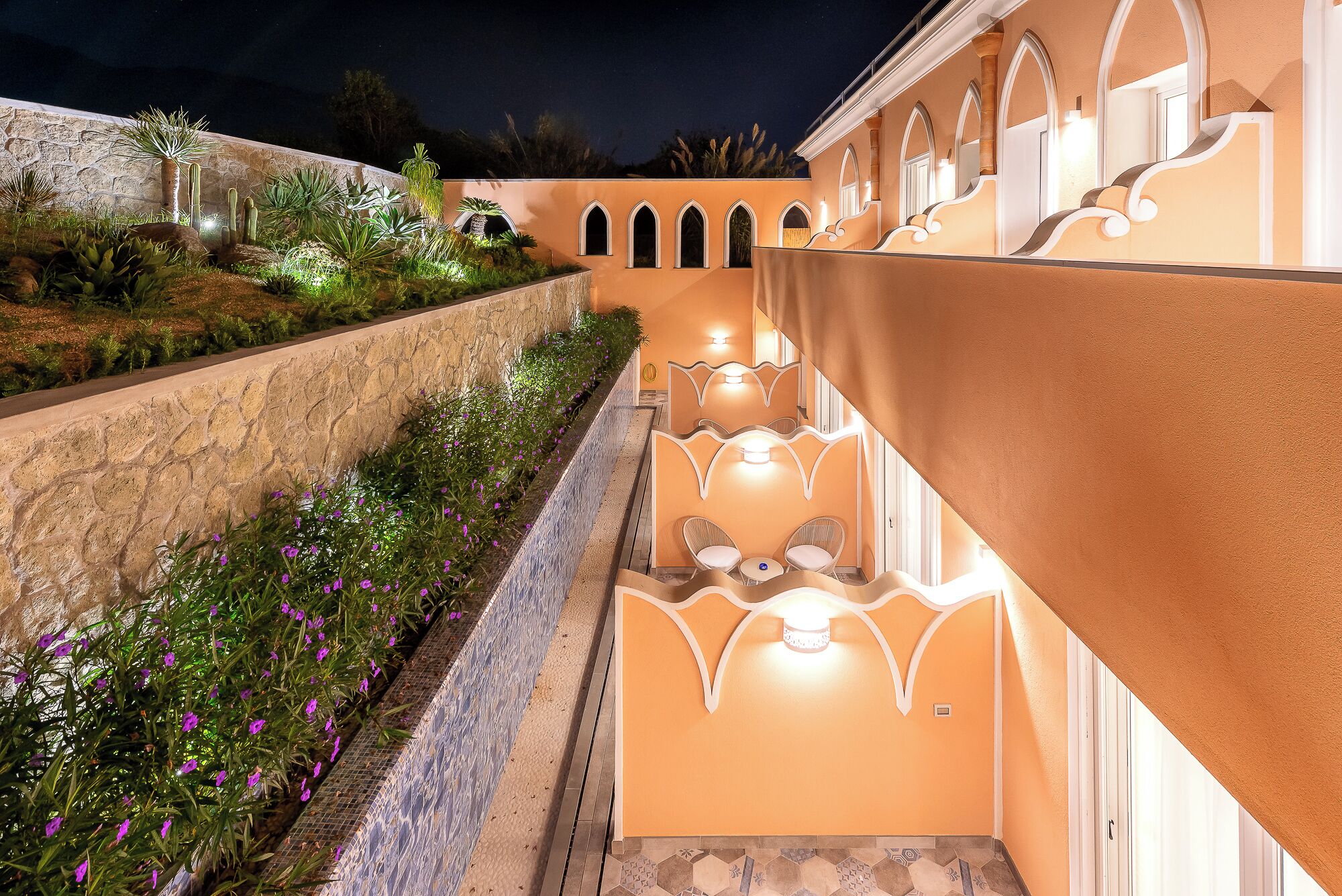 A well-lit outdoor corridor with a stone wall on one side and a building with arched windows on the other.