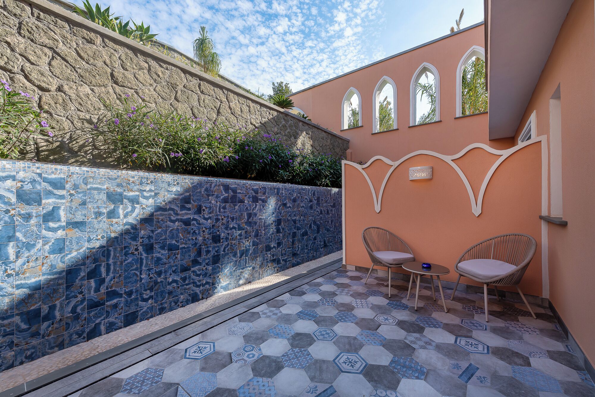Un patio con pared de piedra y suelo de baldosas, dos sillas de mimbre y una lámpara fijada a la pared.