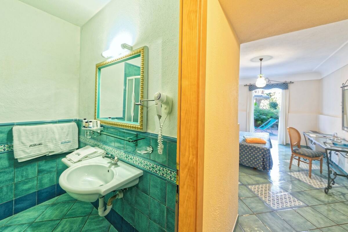 Un baño con lavamanos, espejo y barra para toallas. Hay una puerta que da a un dormitorio con cama y sillón.