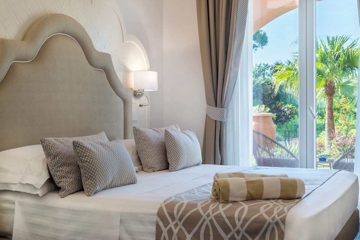 Una cama bien hecha con almohadones estampados, un cabecero con diseño geométrico, una lámpara de noche y vistas a un jardín a través de una ventana.