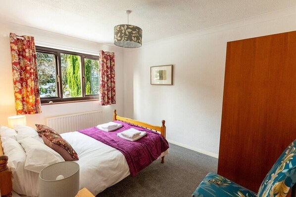 Un dormitorio con cama, una ventana con cortinas, una lámpara de noche, una puerta de madera y un cuadro enmarcado en la pared.