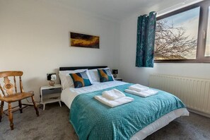 Un dormitorio con cama, una silla, una mesita de noche, un cuadro y una ventana con cortinas.