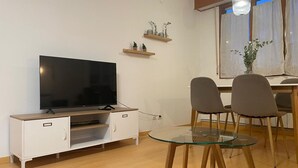 A modern living room with a flat-screen TV on a white cabinet, two grey chairs around a glass table, wooden flooring, and a window with curtains. - Cozy 2BR Apartment with Pool in Jaca – Sleeps 6, 2 Bathrooms (Jaca)