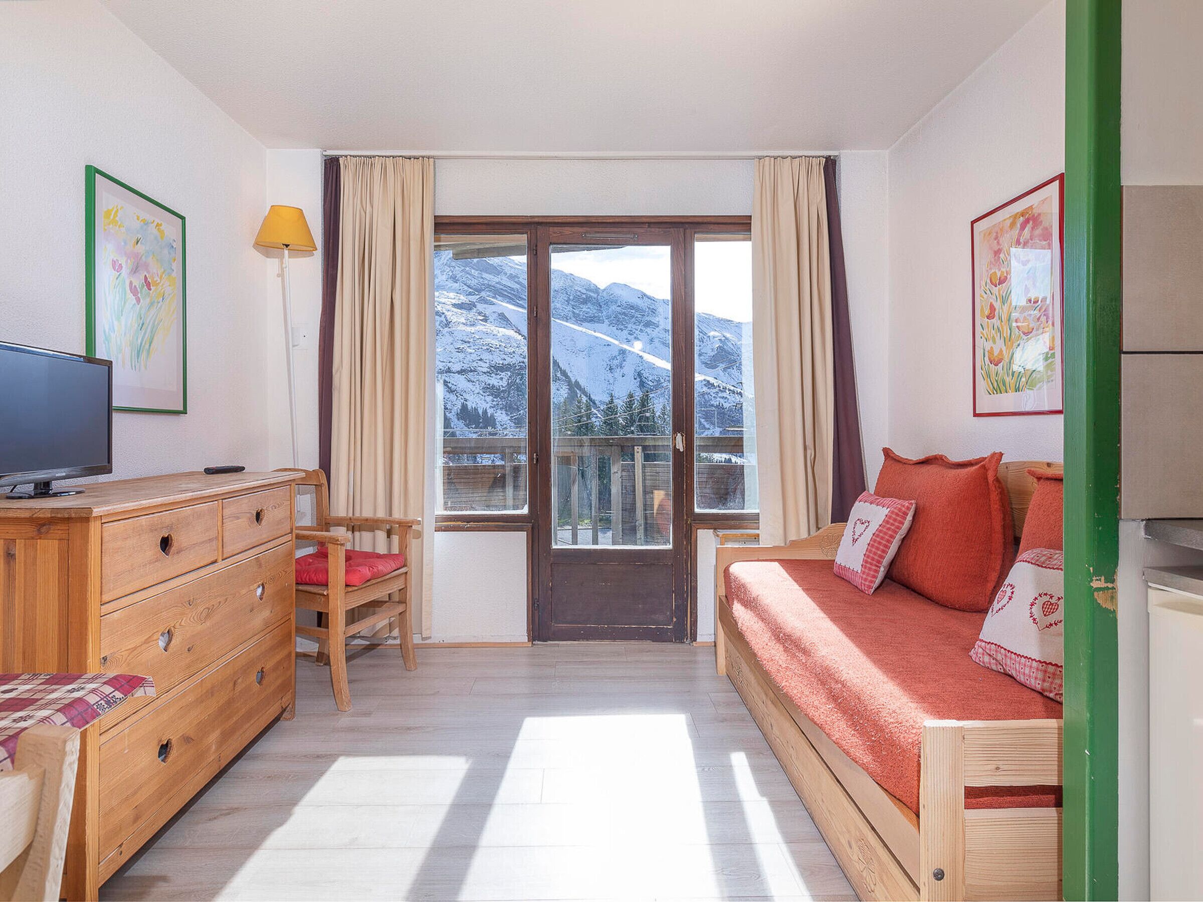 Ett rum med en byrå i trä, en tv, en röd soffa och en utsikt över snöklädda berg genom en glasdörr.