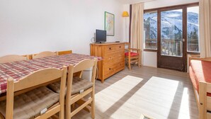 Une pièce chaleureuse avec une table à manger en bois, des chaises, une commode et un téléviseur. Une fenêtre offre une vue sur des montagnes enneigées.