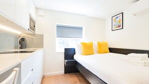 A modern hotel room with a bed, bedside table, and a window with blinds. - StayPrime Fitzrovia Serviced Apartments (London)