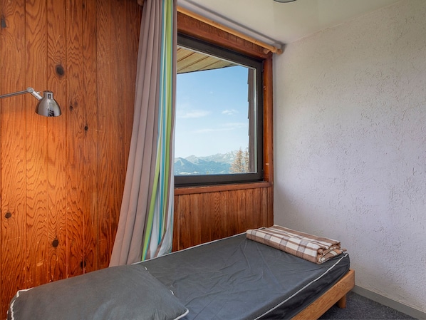 Une petite chambre avec un lit simple, un mur en bois, une fenêtre donnant sur les montagnes et une lampe de chevet.