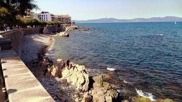 A coastal promenade with a rocky shoreline, a clear view of the sea, and a building in the background.