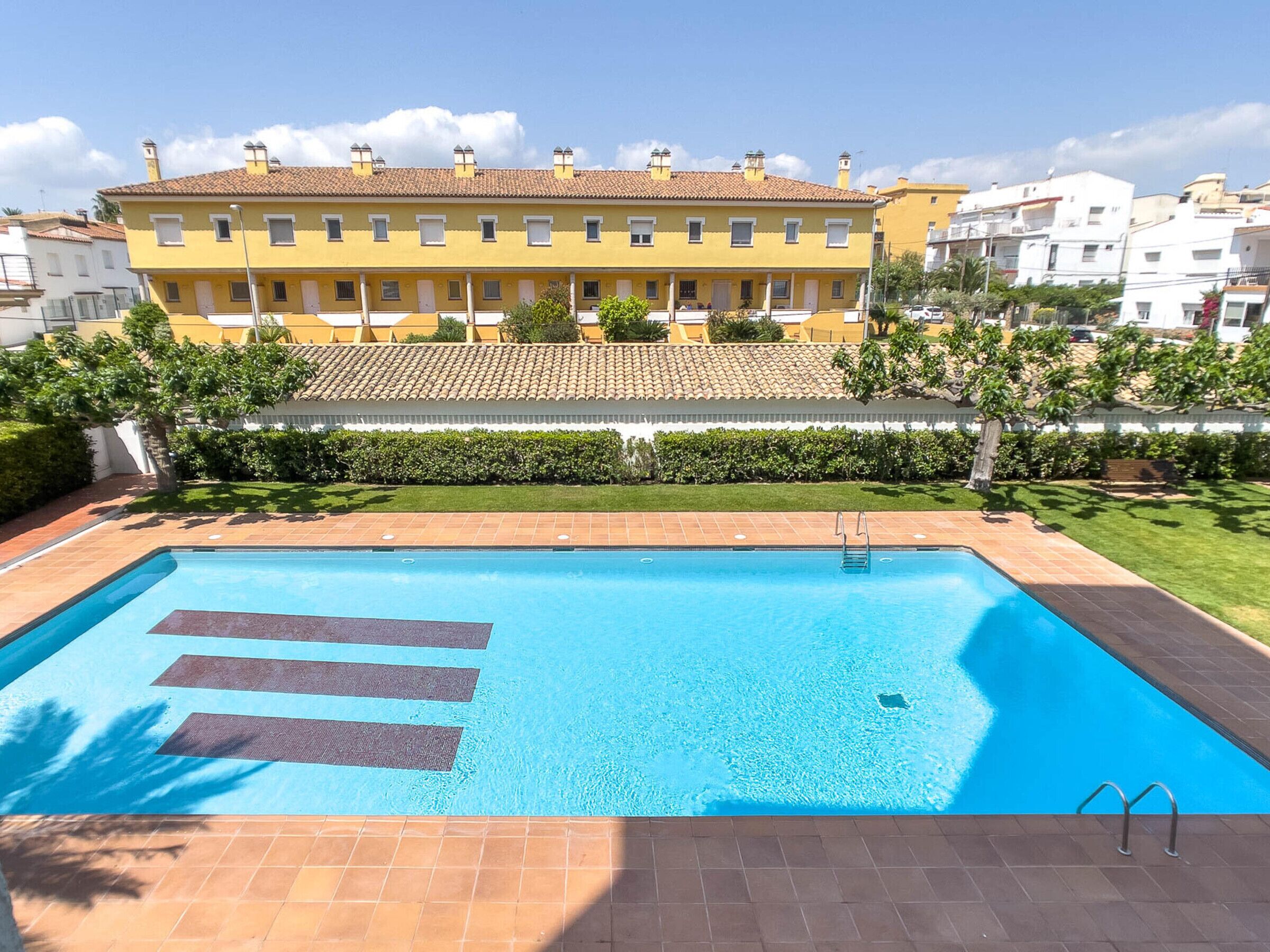 Une piscine rectangulaire à l’eau d’un bleu limpide, entourée d’une terrasse carrelée. Au fond, on aperçoit une rangée de bâtiments jaunes, dotés de nombreuses fenêtres et d’un toit recouvert de tuiles.