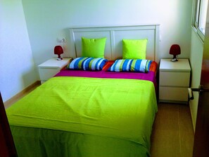 Un lit recouvert d’une couvre-lit vert vif, avec deux oreillers, l’un à rayures et l’autre uni, de couleur verte. De part et d’autre du lit se trouvent deux tables de chevet surmontées de lampes.