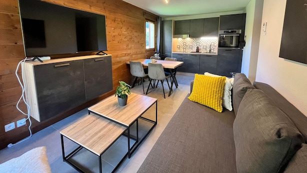 A modern living space with a wooden accent wall, a dining area, a sofa with a yellow pillow, and a kitchen in the background. - Renovated apartment with pool in Samoëns (Samoëns)