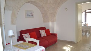 A red sofa, a white chair, a table with a lace tablecloth, a lamp, and a stone archway. - Trulli of the Chain (Martina Franca)