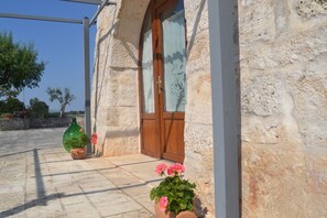 A stone building with a wooden door, a potted plant with pink flowers, and a tiled patio.