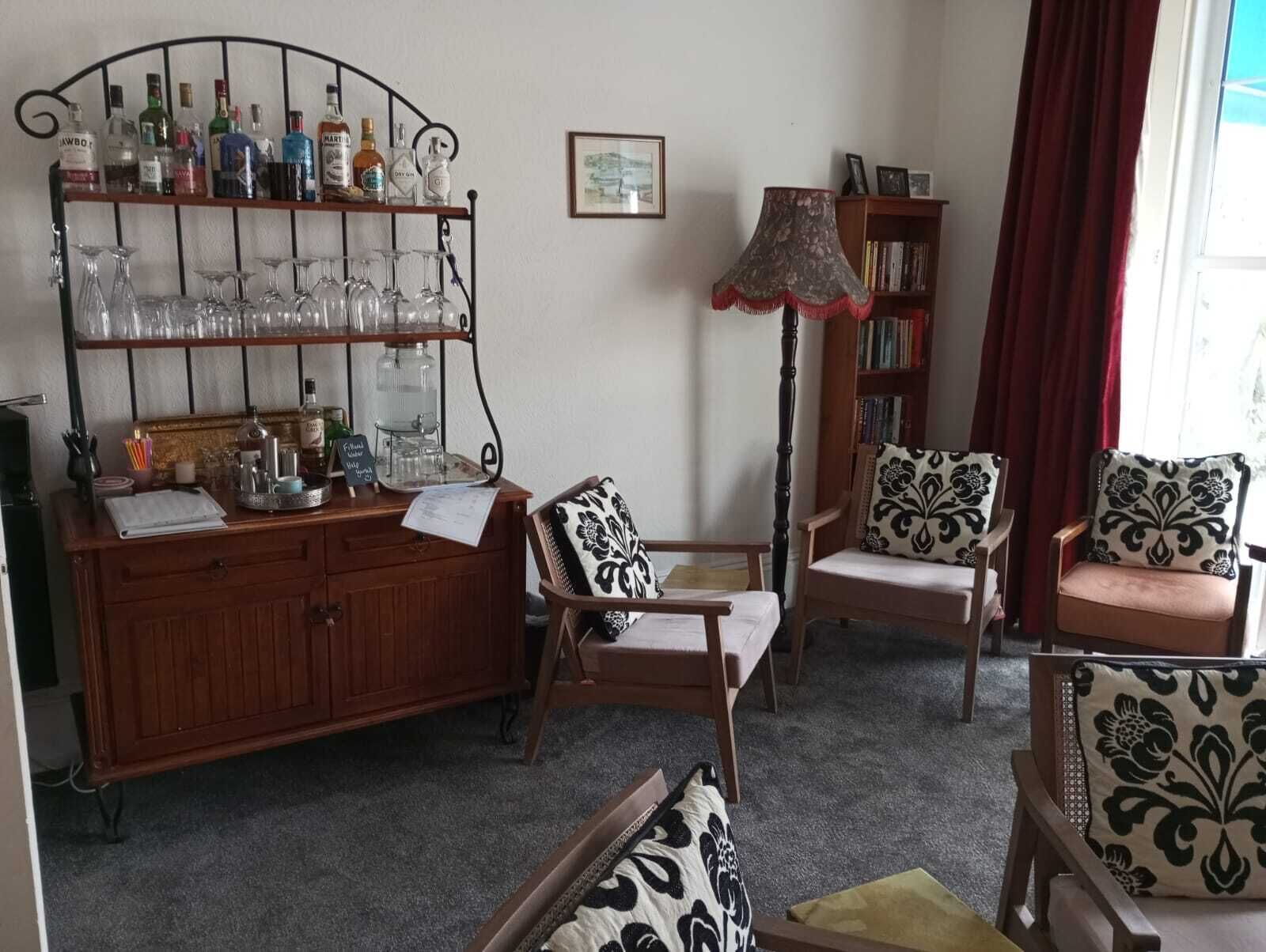 Una sala de estar acogedora con un aparador de madera que exhibe varias botellas y copas, dos sillones estampados, una lámpara de pie y una estantería.