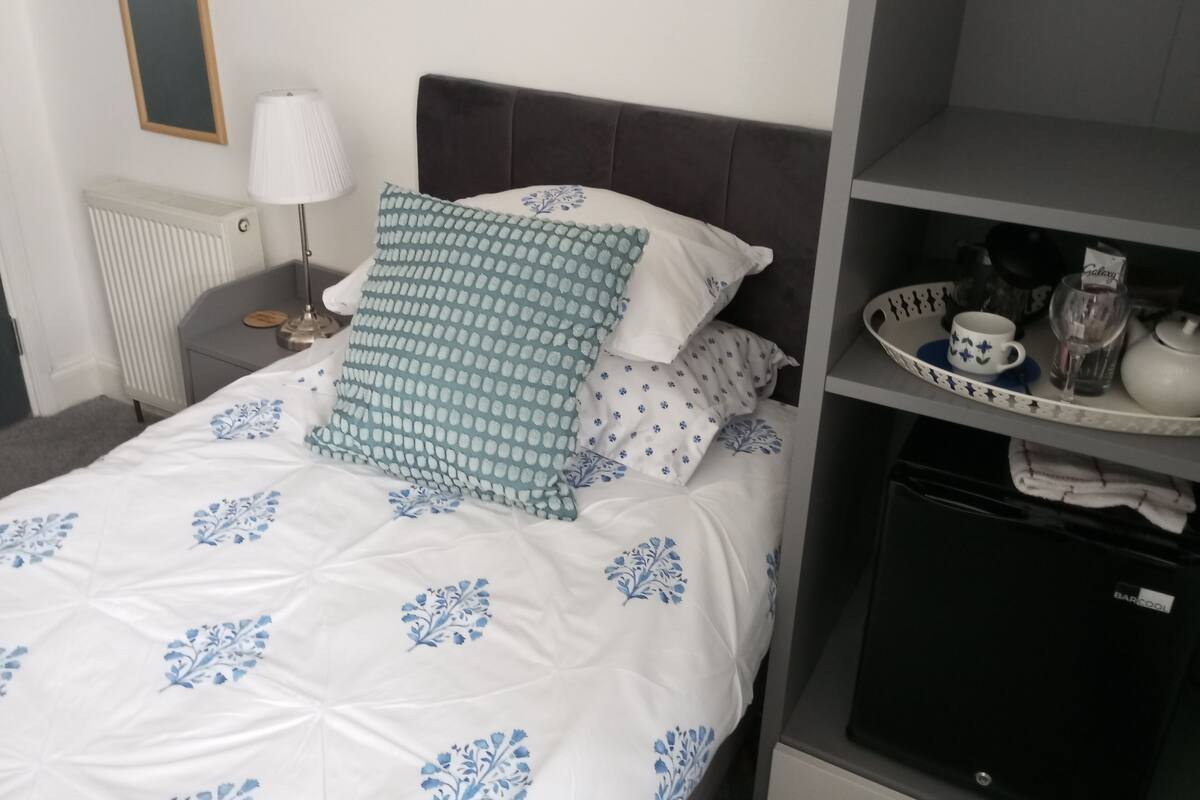 En pænt redt seng med et dynebetræk med blomstermønster, et natbord med en lampe, samt en hylde med te, kop og pynt.
