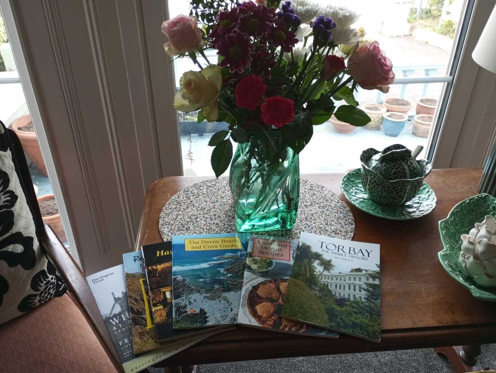 Una mesa con un jarrón de flores, un libro titulado 'The Devon Beach and Cove Guide' y un libro titulado 'TOP BAY'.