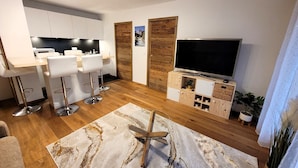 Ein modernes Wohnzimmer mit Holzboden, einem großen Flachbildfernseher, einem Essbereich mit weißen Stühlen und einer Küche mit weißen Schränken.