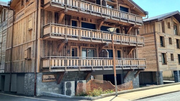 Ein mehrstöckiges Holzhaus mit Balkonen, Stein Fundament und einem Schild mit der Aufschrift 'Le Confidentiel'.