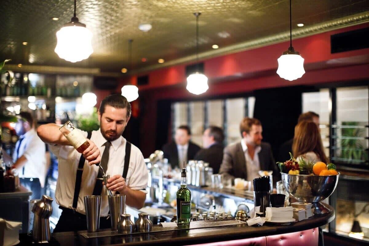 A bar area with a bartender preparing drinks, a bar counter with various bottles and glasses, and a bowl of fruit.