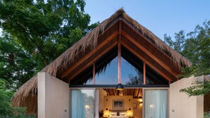 A modern, thatched-roof structure with a large glass door leading to a bedroom with a bed, bedside tables, and a hanging light fixture. - Taru Villas Villu - Willpattu (Eluwankulama)