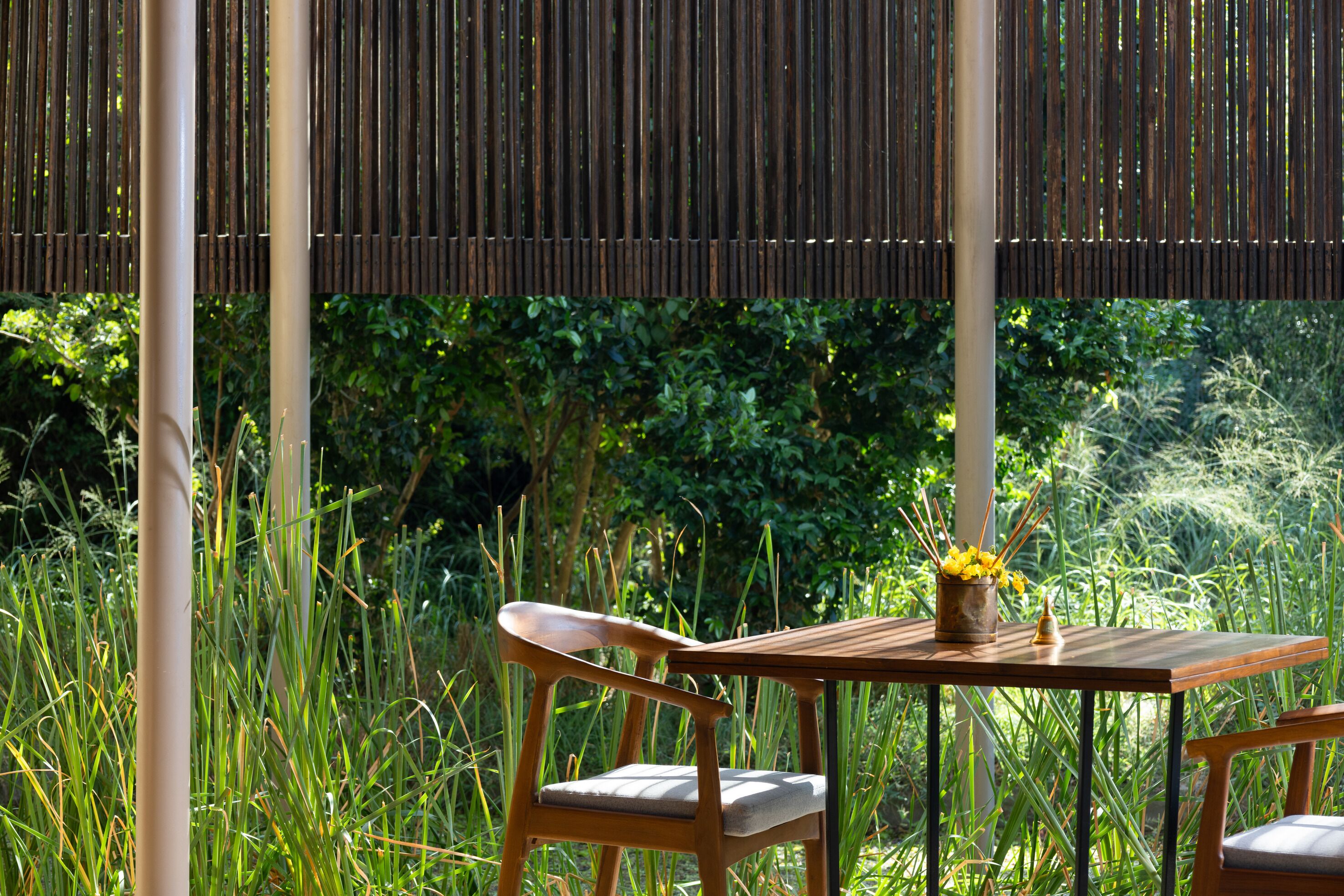 Un conjunto de mesa y sillas de madera en un jardín con valla de bambú y abundante vegetación al fondo.