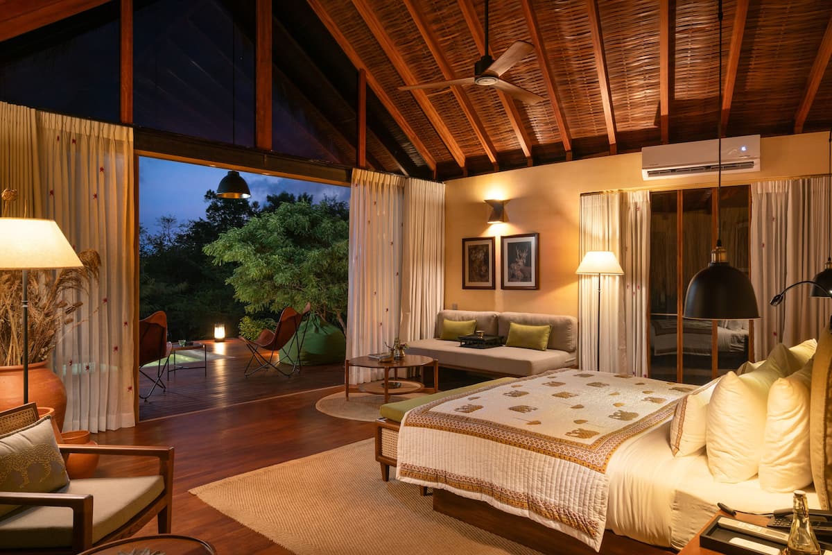 A spacious bedroom with a large bed, a sofa, and a desk. There is a balcony with a view of trees and a lamp.