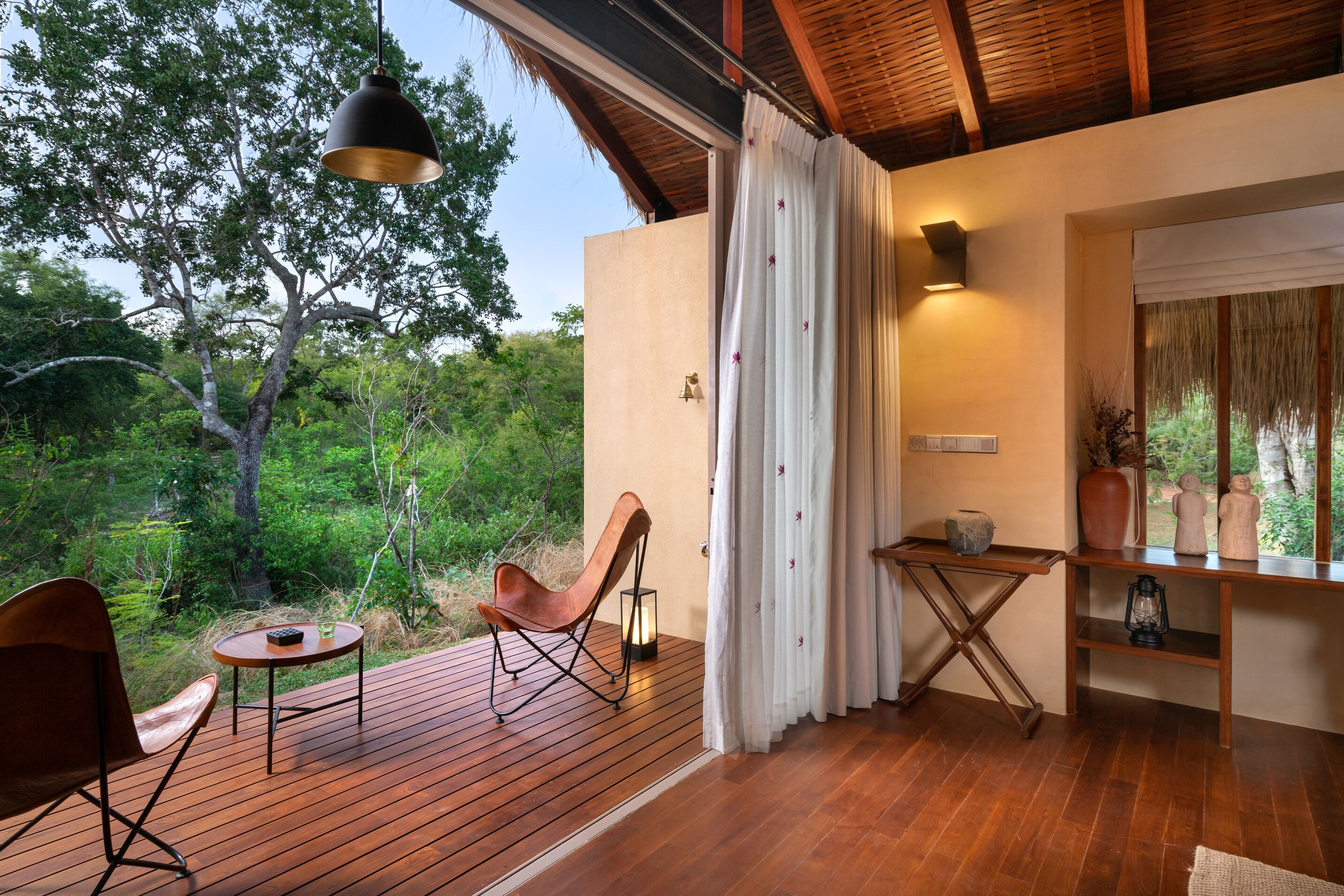Une terrasse en bois avec une chaise et une table, une suspension lumineuse, une table fixée au mur avec une lanterne, et une vue sur les arbres à travers une fenêtre.