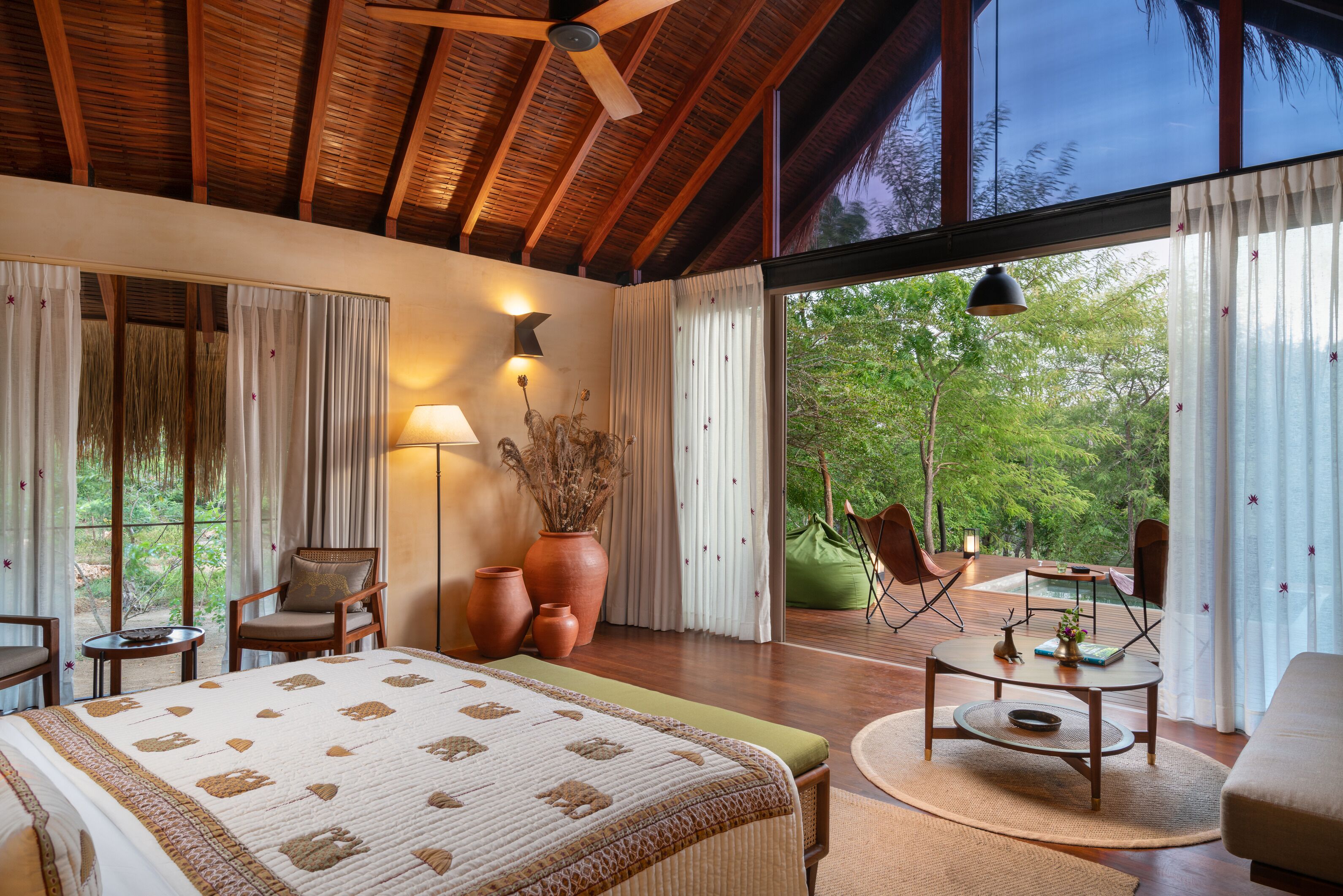 A spacious bedroom with a large bed, a sitting area with a table and chairs, and a view of the outdoors through large windows.