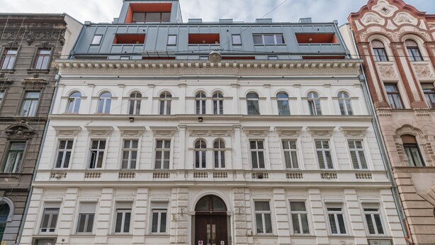 Un immeuble à plusieurs étages doté d’une façade de style classique, avec une entrée centrale surmontée d’un grand portail en arc de cercle et de nombreuses fenêtres.